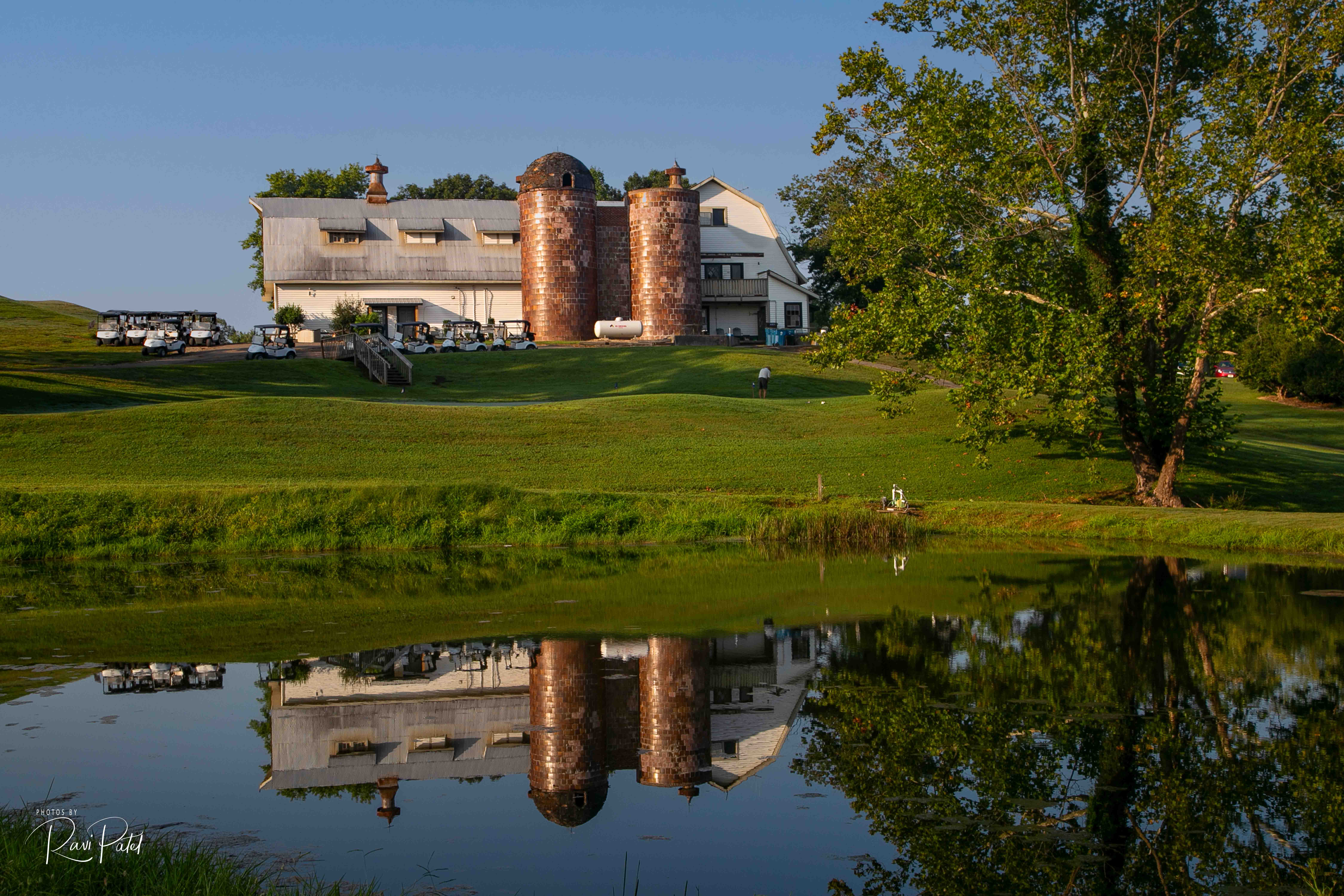 View from the lake. The Silos Golf Club clubhouse in the background with it's reflection visible on the lake's surface. A tree sits on the right and the sky is clear blue.