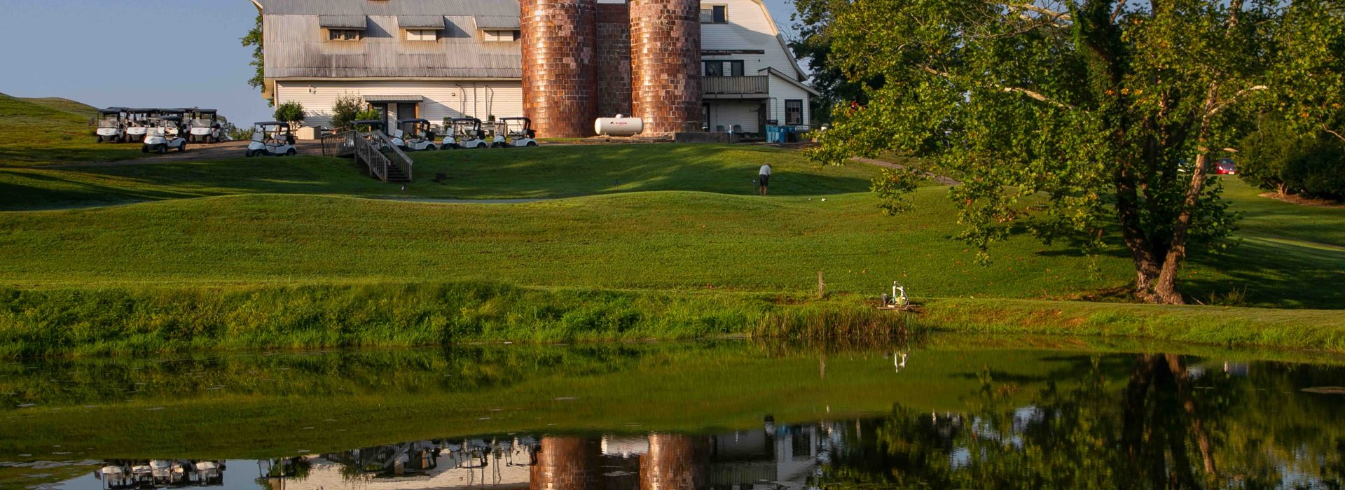 View from the lake. The Silos Golf Club clubhouse in the background with it's reflection visible on the lake's surface. A tree sits on the right and the sky is clear blue.