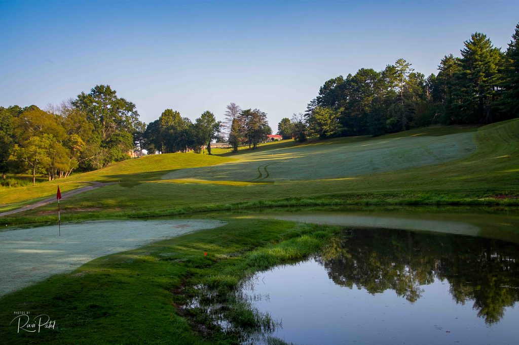 A lake to the right and a golf course hole to the left. A red flag marks the spot. Clear blue skies and a line of trees shapes the backgrond.